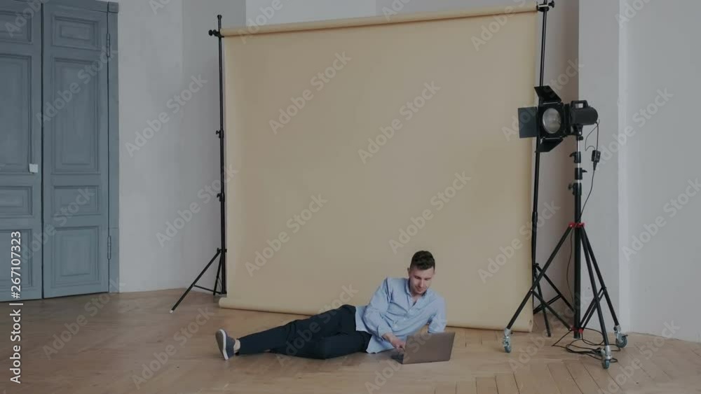 Young handsome male videoblogger working with laptop in comfort position while lying on the floor. He editing video for his video blog chanel. Freelance or remote work indoor.