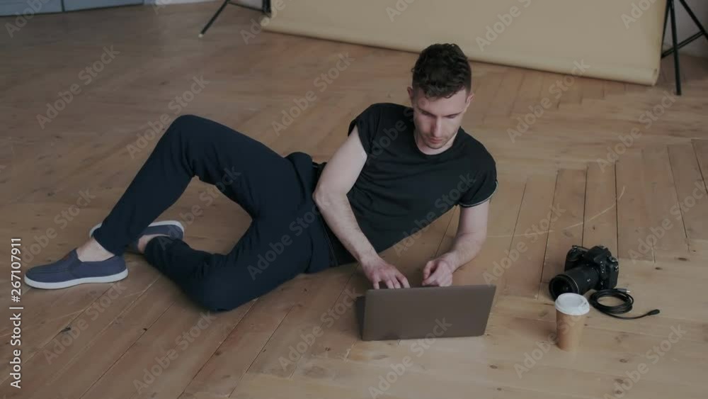 Young handsome male videographer wearing black shirt and trousers. He working with laptop in comfort position while lying on the floor. He editing video for his video blog chanel.