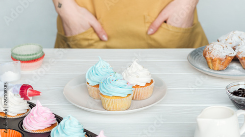 Cropped view of woman with hands in pockets near table with cupcakes isolated on grey