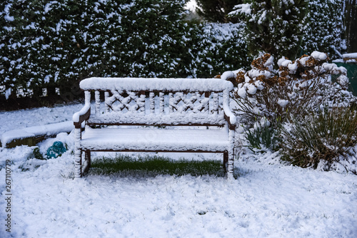 Snow filled Winter landscape of wood vintage bench with white snowy coating in Welsh garden on freezing cold day with lawn and plants.