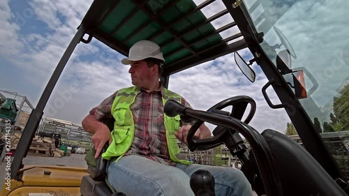 Caucasian man, storehouse worker dressed in a reflective vest and white helmet working on a forklift outdoors. Action camera shot, wide angle.
