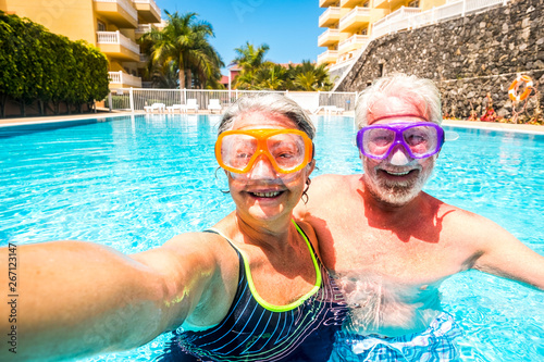 Happy cheerful people old senior man and woman have fun together in the summer swimming pool activity taking selfie pictures with scuba masks on the face for funny outdoor leisure activity in vacation © simona
