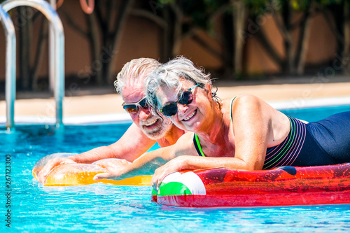 People retired old senior man and woman couple enjoying summer swimming pool leisure activity lay down on coloured trendy fashion lilos inflatable mattress on blue water at home or hotel activity photo