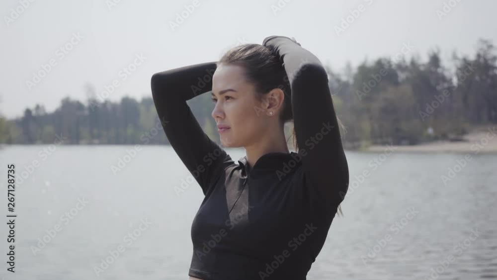 Attractive slim girl straightens her horsetail, standing with arms up in front of the river. Beautiful landscape in the background. Leisure outdoors Attractive slim girl straightens her horsetail, standing with arms up in front of the river. Beautiful landscape in the background. Leisure outdoors