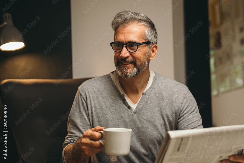 portrait of man sitting sofa reading newspaper and drinking coffee at his home portrait of man sitting sofa reading newspaper and drinking coffee at his home