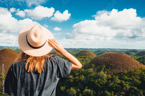 Woman in straw hat standing in front of the Chocolate Hills in the Bohol island in the Philippines. Hills covered in brown grass. Famous touristic place