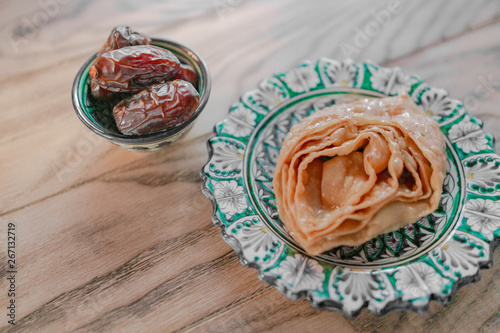Traditional oriental sweetness baklava, puff pastry confection in syrup, in Eastern plate with a national pattern. Date fruits in the bowl near. Horizontal image. Wooden background.