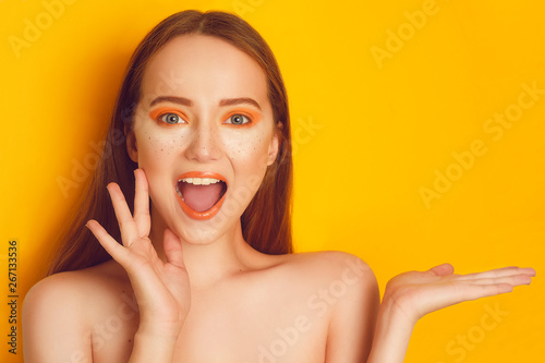 Beautiful girl with shiny brown straight long hair. Woman with orange makeup with freckles. Girl near empty copy space shows product . Girl surprised, on bright orange background