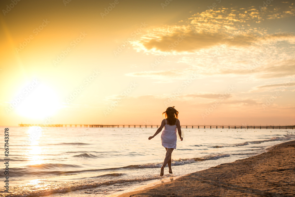 Slim young woman in white dress running on the beach at summer sunset