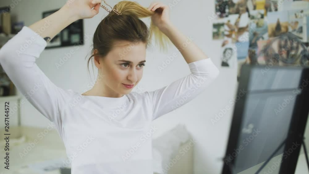 Woman is looking at the mirros and tying her long smooth hair in ponytail, making hairdo Woman is looking at the mirros and tying her long smooth hair in ponytail, making hairdo