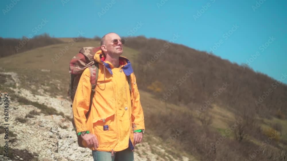 A man in a yellow jacket, glasses and a large tourist backpack stands in the mountains, enjoying the scenery. In the background are hills and sky. Mountain landscape. A man in a yellow jacket, glasses and a large tourist backpack stands in the mountains, enjoying the scenery. In the background are hills and sky. Mountain landscape.
