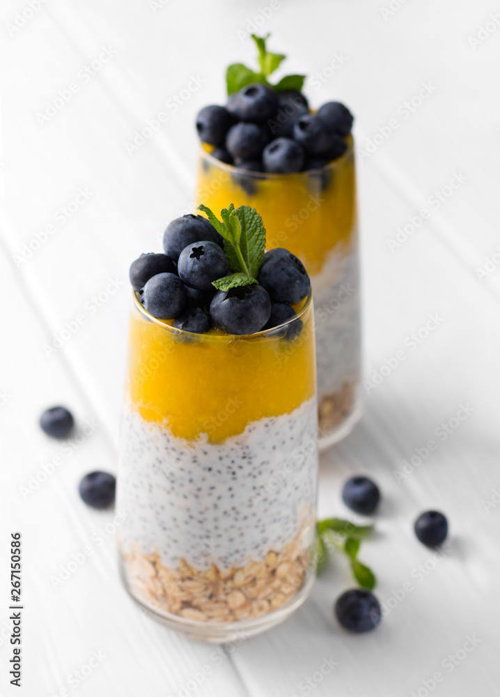 Glass of yogurt with chia seed, blueberries and mango mousse and oatmeal on a white wooden table. Glass of yogurt with chia seed, blueberries and mango mousse and oatmeal on a white wooden table.