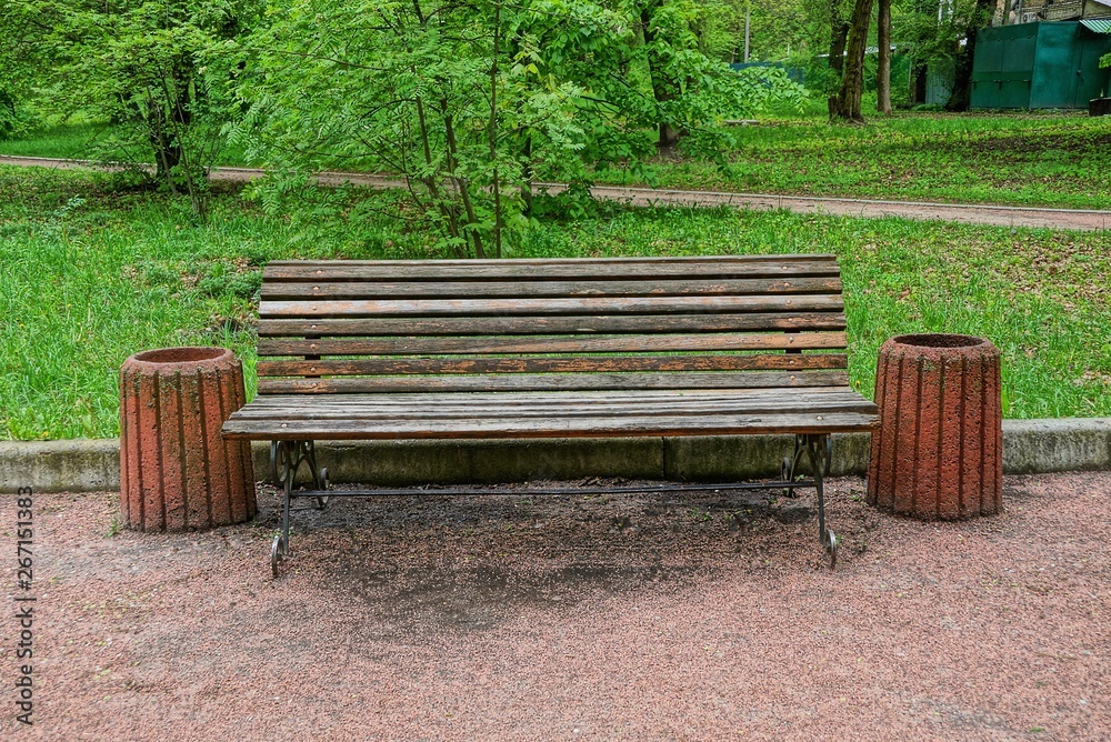 brown wooden bench and a urn are standing on an alley in the park among green grass and trees brown wooden bench and a urn are standing on an alley in the park among green grass and trees