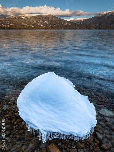 A rock covered in snow and ice sits on the shore of Priest Lake Idaho. Mountains glow in the setting sun in the background.