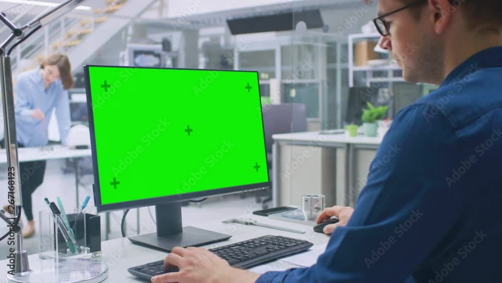 Over the Shoulder Shot of Engineer Working with Green Mock-up Screen Desktop Computer. In the Background Engineering Facility with Specialists Working on Blueprints and Drawings with Industrial Design