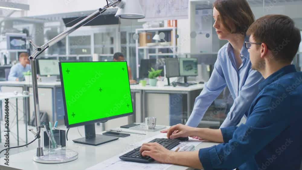 Engineer Working with Green Mock-up Screen Desktop Computer Is advised by Female Project Manager. Engineering Facility with Specialists Working on Blueprints and Drawings with Industrial Design