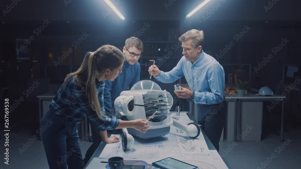 Late at Night in Robotics Engineering Facility Three Technical Engineers Talk and Work on a Wheeled Robot Prototype. In the Background High Tech Research Center with Screens Showing Industrial Design Late at Night in Robotics Engineering Facility Three Technical Engineers Talk and Work on a Wheeled Robot Prototype. In the Background High Tech Research Center with Screens Showing Industrial Design