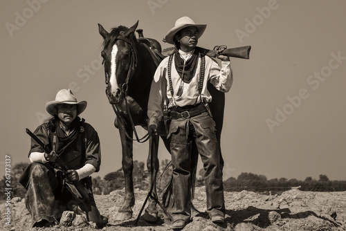 Vintage photo, of two men wearing a cowboy outfit with a horse and a gun held in the hand.