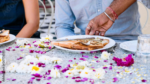 Waiter brings breakfast toasts and pancakes to a couple sitting at the table covered with flowers petals in hotel. Table close-up.