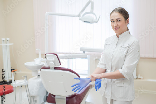 Attractive female doctor in white uniform puts on gloves. Young beautiful female dentist brunette in the office by the chair wearing gloves.