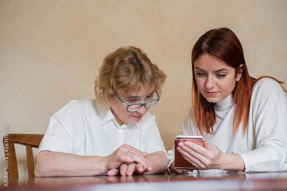 Indoor image of a beautiful red-haired young woman holding a mobile phone, showing her attractive elderly mother how to use an electronic gadget. Girl show how to use the smartphone to her mother. Indoor image of a beautiful red-haired young woman holding a mobile phone, showing her attractive elderly mother how to use an electronic gadget. Girl show how to use the smartphone to her mother.