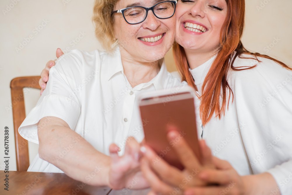 An elderly mother and daughter dressed in light clothes are smiling and taking selfies. Mom and daughter are photographed on the front camera of the smartphone while sitting at home at the table. An elderly mother and daughter dressed in light clothes are smiling and taking selfies. Mom and daughter are photographed on the front camera of the smartphone while sitting at home at the table.