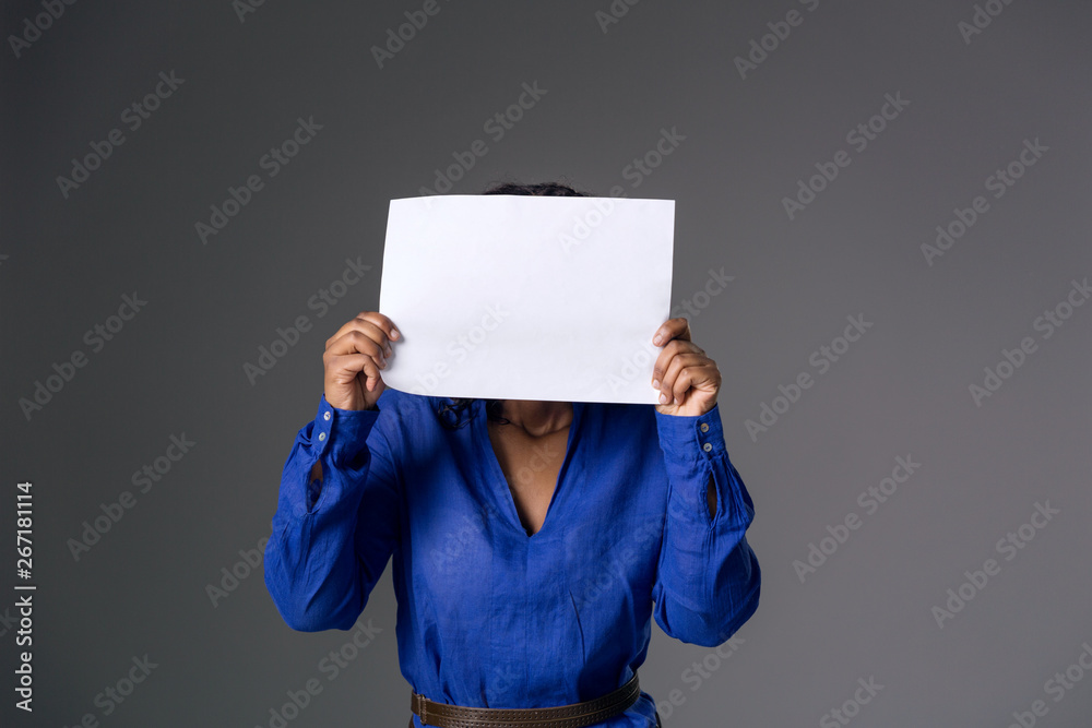 A dark-skinned girl in a blue blouse covered her face with a white sheet of paper on a gray background. A dark-skinned girl in a blue blouse covered her face with a white sheet of paper on a gray background.