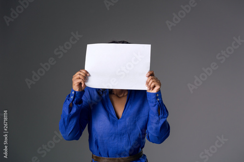 A dark-skinned girl in a blue blouse covered her face with a white sheet of paper on a gray background.