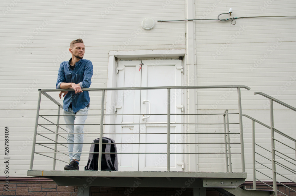 bearded young man in jeans and denim shirt is standing on porch of industrial building near closed door. job search concept