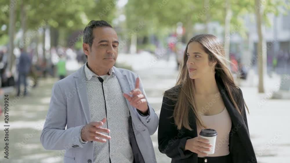 Male and female colleagues talking outdoor. Handsome mature man and smiling young woman with coffee to go walking on street and talking together. Communication concept Male and female colleagues talking outdoor. Handsome mature man and smiling young woman with coffee to go walking on street and talking together. Communication concept