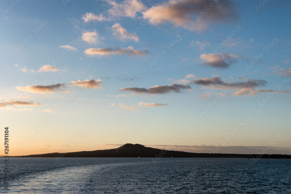Silhouette of Rangitoto Island, Auckland, New Zealand over the ocean, with a dramatic Sunet/Sunrise and a boat wake traversing the image