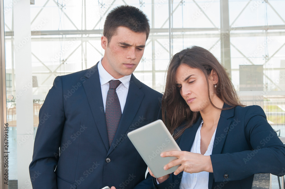 Pretty serious woman in white blouse and concentrated young man in dark suite standing in office corridor, woman showing data on tablet. Work, communication concept Pretty serious woman in white blouse and concentrated young man in dark suite standing in office corridor, woman showing data on tablet. Work, communication concept
