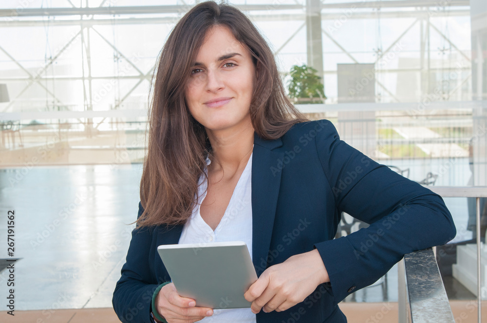 Pretty smiling Caucasian woman in white blouse and navy blue jacket standing in office corridor, looking at camera, holding tablet in hands. Work concept Pretty smiling Caucasian woman in white blouse and navy blue jacket standing in office corridor, looking at camera, holding tablet in hands. Work concept