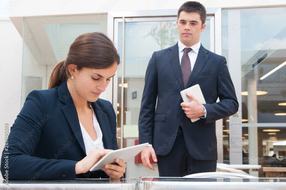 Focused business woman using tablet and coworker standing near. Business woman sitting at desk and man standing and holding tablet computer. Business people and technology concept. Focused business woman using tablet and coworker standing near. Business woman sitting at desk and man standing and holding tablet computer. Business people and technology concept.