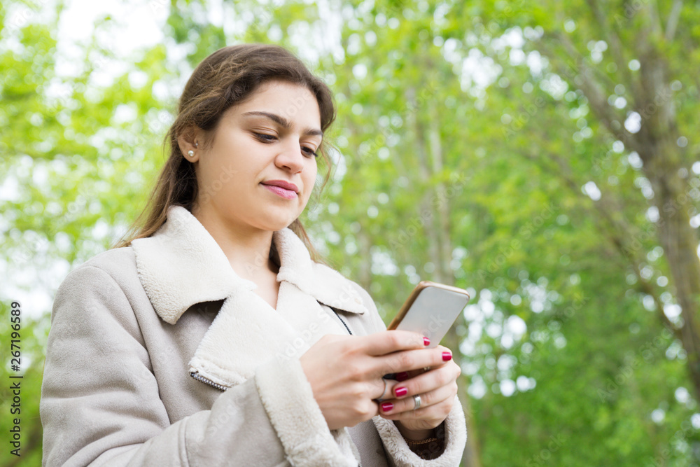 Content pretty young woman using smartphone in park. Lady wearing jacket, texting on gadget and standing with green trees in background. Communication and nature concept. Content pretty young woman using smartphone in park. Lady wearing jacket, texting on gadget and standing with green trees in background. Communication and nature concept.