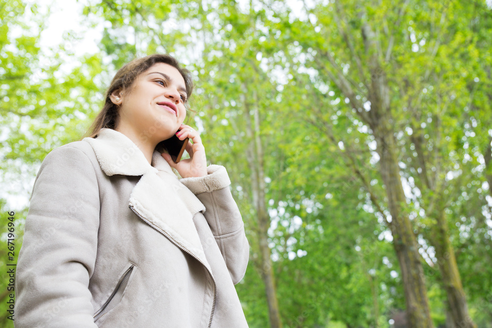 Smiling pretty young woman calling on smartphone in park. Lady wearing jacket, talking on mobile phone and standing with green trees in background. Communication and nature concept. Smiling pretty young woman calling on smartphone in park. Lady wearing jacket, talking on mobile phone and standing with green trees in background. Communication and nature concept.