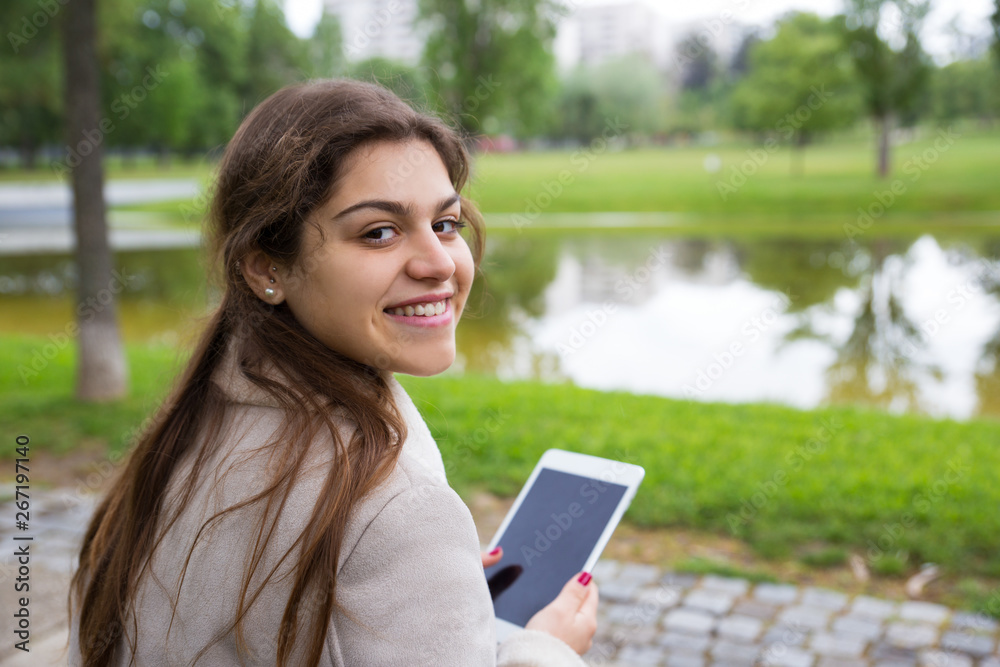 Happy joyful college girl working on class project in park. Young woman in warm jacket sitting on bench outdoors and using tablet. Digital technology concept Happy joyful college girl working on class project in park. Young woman in warm jacket sitting on bench outdoors and using tablet. Digital technology concept