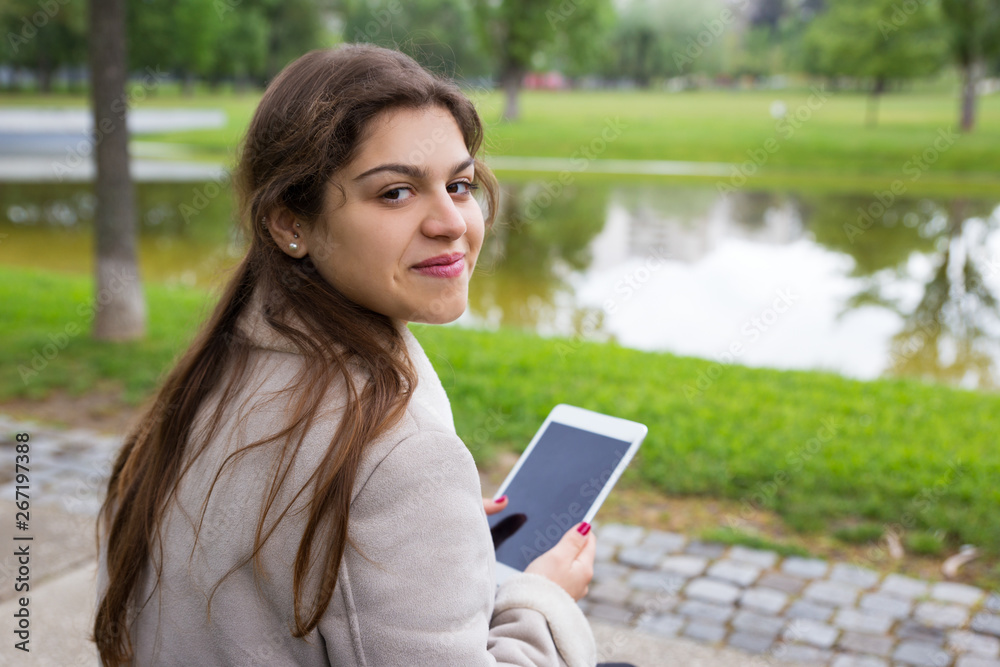 Positive Latin girl chatting online outdoors. Back view of young woman sitting on bench in park, holding tablet, turning around and smiling at camera. Communication concept Positive Latin girl chatting online outdoors. Back view of young woman sitting on bench in park, holding tablet, turning around and smiling at camera. Communication concept