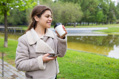 Smiling relaxed girl with phone drinking tasty coffee in park. Young woman in warm casual jacket standing near pond, holding smartphone and enjoying takeaway drink. Outdoor leisure concept
