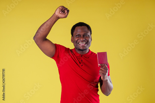 handsome happy young african man Kenya Africa tourist isolated yellow wall background holding tickets with passport and coctail glass trip around the world