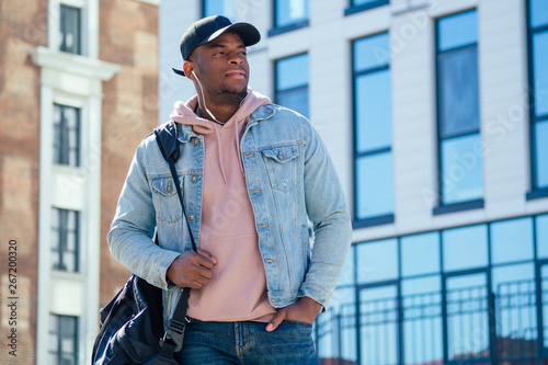Street fashion concept - stylish handsome afro american man in a pink shirt hood and denim jacket and black cap against the windows of the business center