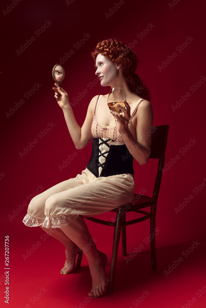 Medieval redhead young woman as a duchess in black corset and night clothes sitting on red background with a mirror and a glass of wine. Concept of comparison of eras, modernity and renaissance.