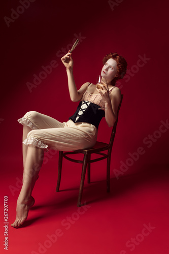 Medieval redhead young woman as a duchess in black corset and night clothes sitting on red background with a mirror and a glass of wine. Concept of comparison of eras, modernity and renaissance.