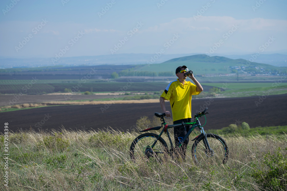 A man on a bicycle stopped on a hill and drinks water.