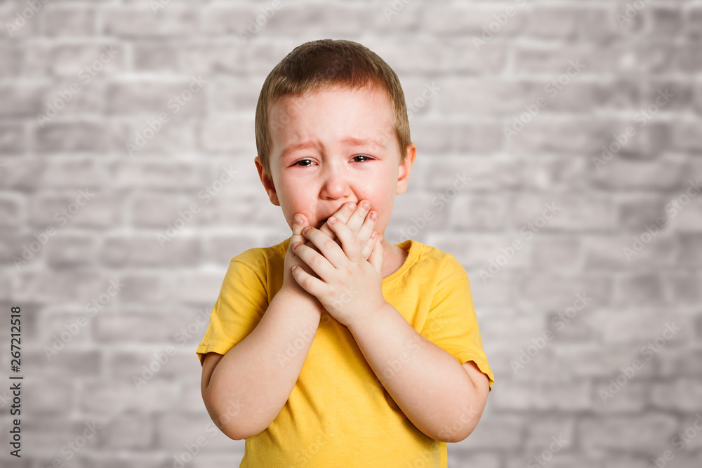 Crying baby boy in a yellow T shirt covers his face with hands and shouts, studio on brick wall background Crying baby boy in a yellow T shirt covers his face with hands and shouts, studio on brick wall background