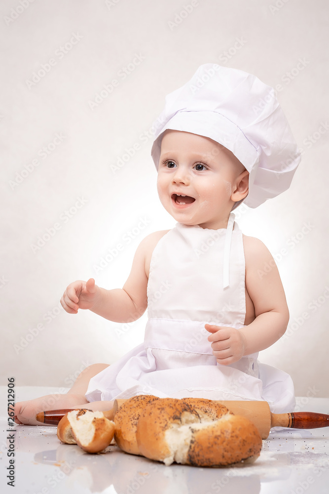 Charming toddler baby in hat of cook and apron sitting with bread loaves, laughing happily Charming toddler baby in hat of cook and apron sitting with bread loaves, laughing happily