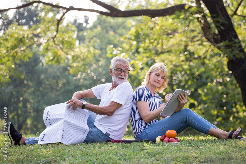 Fototapeta premium Happy senior couple relaxing sitting back to back in park reading newspaper and take write notes to Diary book to together . old people in the summer park . Elderly resting .mature relationships .