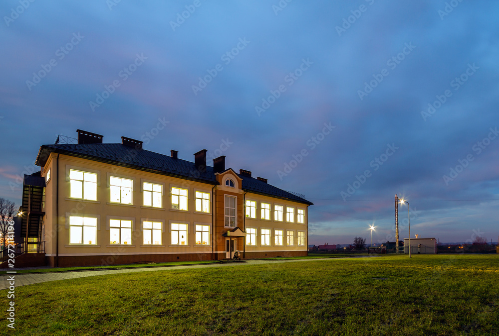 New modern two-storied kindergarten preschool building with brightly lit windows on green grassy lawn and evening blue sky copy space background. Architecture and development concept. New modern two-storied kindergarten preschool building with brightly lit windows on green grassy lawn and evening blue sky copy space background. Architecture and development concept.