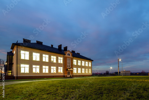 New modern two-storied kindergarten preschool building with brightly lit windows on green grassy lawn and evening blue sky copy space background. Architecture and development concept.