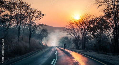 Cyclist passing fire and smoke with the sun reflecting on tarmac during sunset, on mountainbike riding in Zambian mountains,